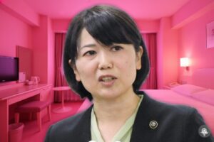 複数のマイクを前に、黒いジャケット姿で話す小川晶前橋市長。背景は、ベッドやカーテンが置かれ、全体がピンク色の照明で照らされたホテルの一室に差し替えられている。